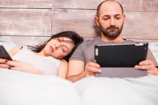 Beautiful wife in pajamas sleeping while her husband is working on tablet computer.