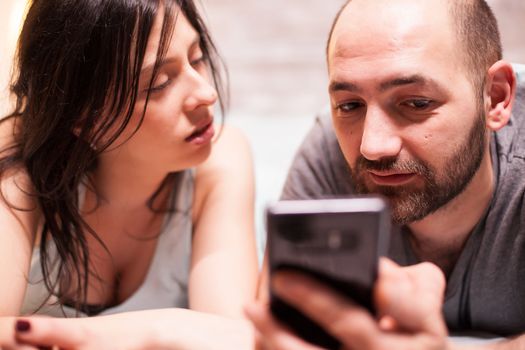 Close up of young man in pajamas using smartphone while his wife is looking at him.
