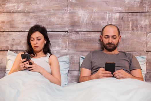 Caucasian young couple under the blanket wearing pajamas using smartphone.