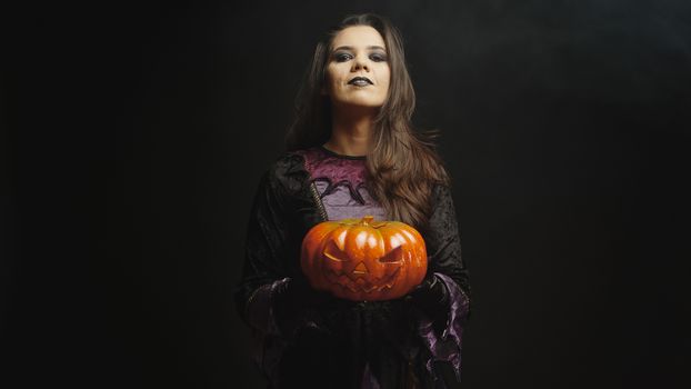 Beautiful young woman with evil face dressed up like a witch holding a pumpkin for halloween over a black background