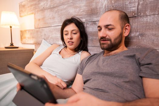 Beautiful married couple in pajamas lying in bed watching tv show on tablet computer.