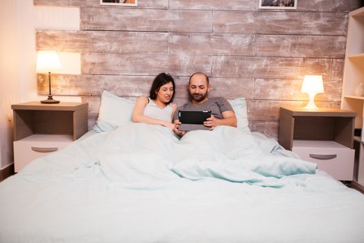 Married caucasian couple wearing pajamas using tablet computer before sleep.