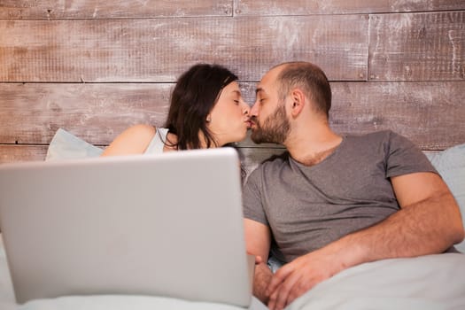Couple in pajamas kissing laying in bed while using laptop.