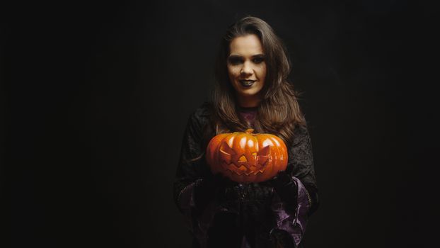 Young woman dressed up like a witch holding a pumpkin for halloween looking at the camera over a black background.