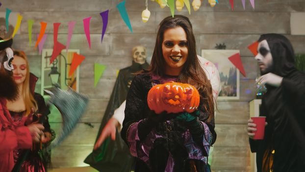 Beautiful young woman dressed up like a scary witch with a pumpkin at a halloween party with her friends.