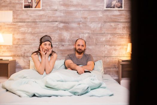 Beautiful wife in pajamas watching a shocking movie on tv together with her bored husband.