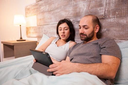 Married young couple wearing pajamas using tablet computer to plan their vacation before bedtime.