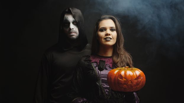 Young woman dressed up like a witch holding a Jack o Lantern for Hallowee next to a dark grim reaper over a black background