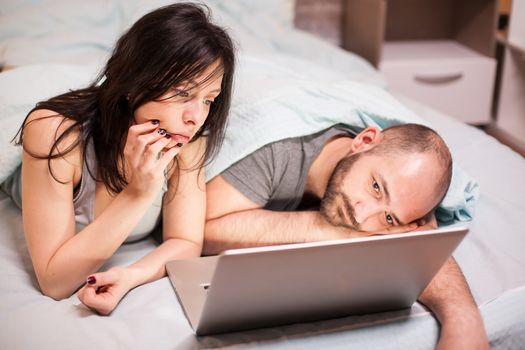 Bored young couple in pajamas watching a movie on laptop.