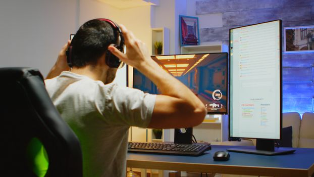 Back view of man with headphones playing shooter game sitting on gaming chair. There are two streaming chat opened on the playing session