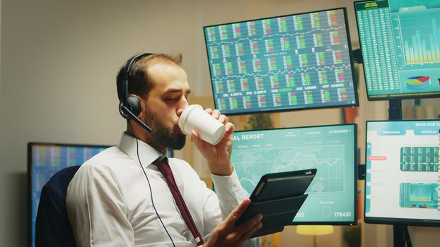 Stock market trader having a conversation on headphones while using tablet computer and taking a sip of coffee in home office.