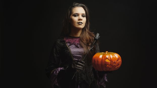 Young woman dressed up like a witch for halloween holding a scary pumpkin over black background