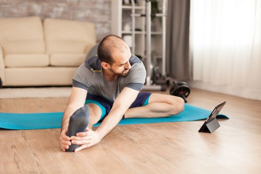 Fit man at home watching online stretching lesson on tablet computer during covid-19 epidemic.