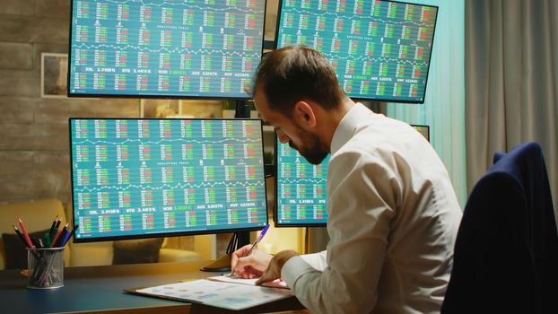 Bearded businessman taking notes while checking stock market from home office.