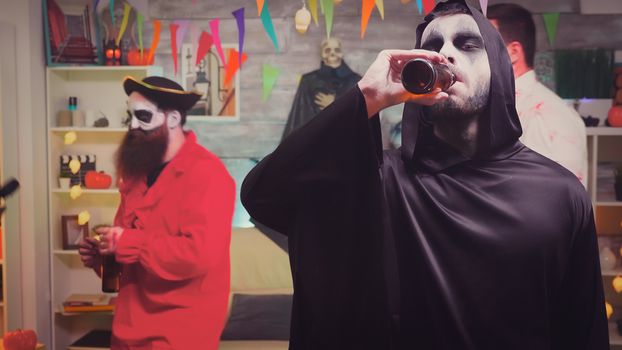 Zoom in shot of man dressed up like grim reaper drinking beer at halloween party.