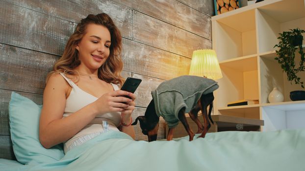 Attractive woman in pajamas browsing on her smartphone lying in bed with her happy dog.