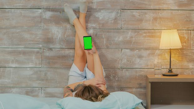 Back view of woman wearing pajamas lying in bed using phone with green screen.