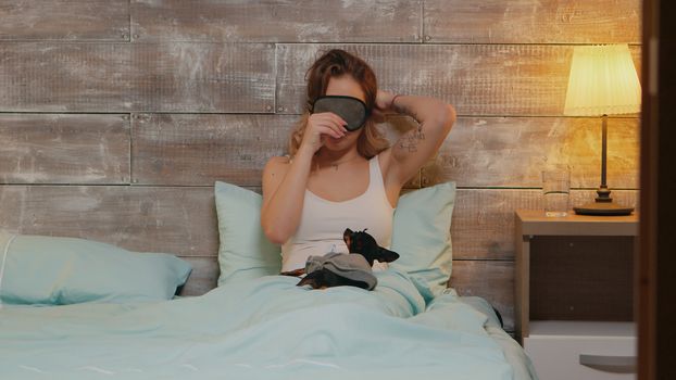 Beautiful young woman in pajamas playing with her dog before using sleep mask for sleeping.