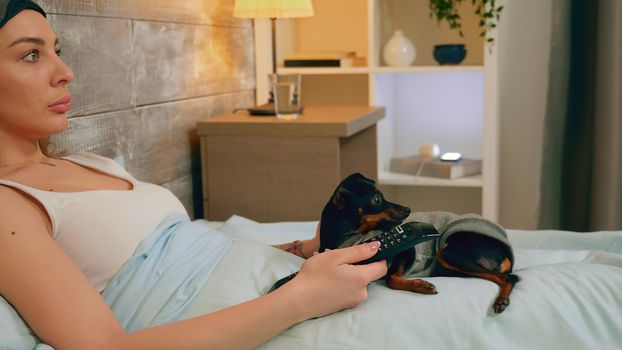 Close up woman in pajamas and her dog watching tv in bed.