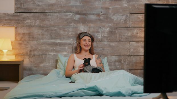 Beautiful woman in pajamas with sleep mask watching tv with her dog.