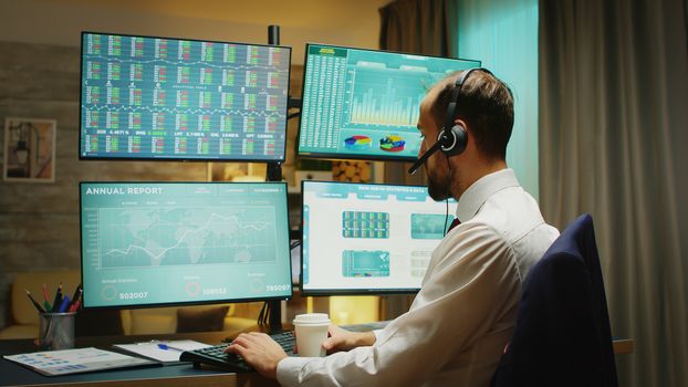 Businessman in home office drinking coffee and talking on headphones about financial crisis. Four big displays with different stock market data