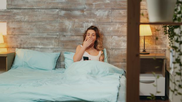 Beautiful woman in pajamas yawning while using smartphone before bedtime.