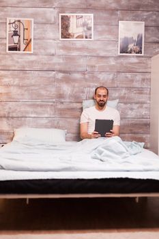Smiling man browsing on tablet computer wearing pajamas and laying in bed at night.
