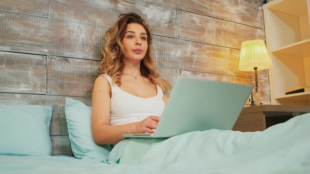Caucasian woman in pajamas reading an online article on her laptop while resting in the bed