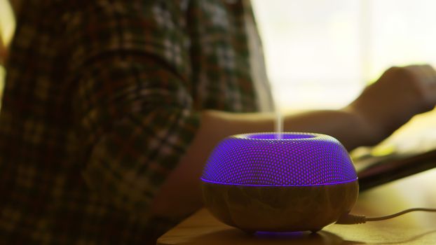 Mist from blue led electric essential oil diffuser while woman is working on laptop.