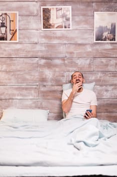 Tired man laying in comfortable bed yawning wearing pajamas and using smartphone.
