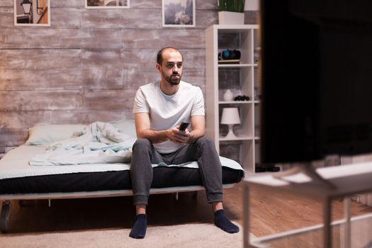 Man sitting on the edge of his bed at night watching tv holding remote control.