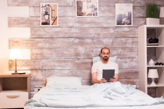 Cheerful man browsing on tablet computer laying in bed at night.