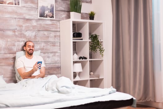 Cheerful man in pajamas laughing while watching a funny video on smartphone at night in comfortable bed.