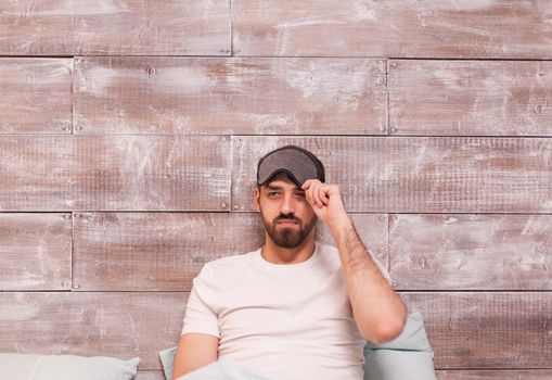 Tired man in comfortable bed pulling his sleep mask over eyes.