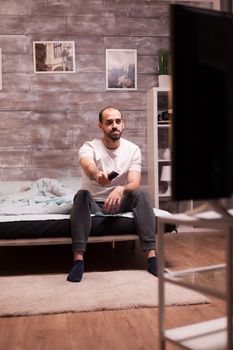 Man sitting on the edge of his bed at night changing channels on tv with remote control.