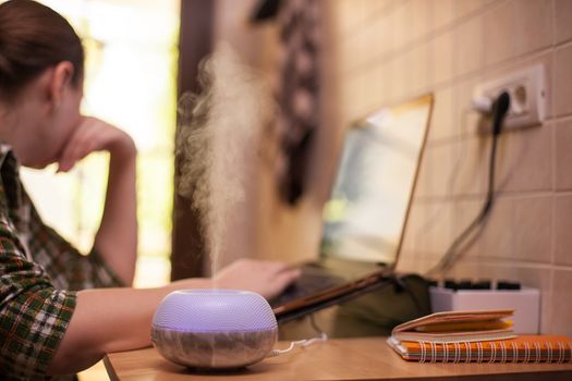 Mist coming out of essential oil diffuser with purple led while woman working on laptop.