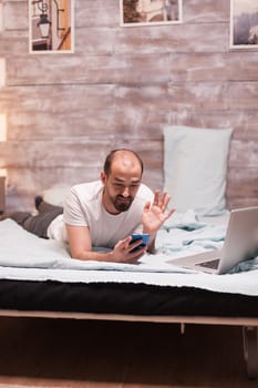 Man at night in bed during a video call waving laying in comfortable bed.