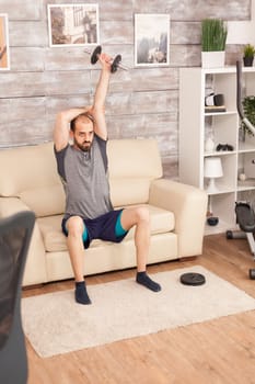 Athletic man training triceps with dumbbell sitting on sofa during covid-19 self isolation.