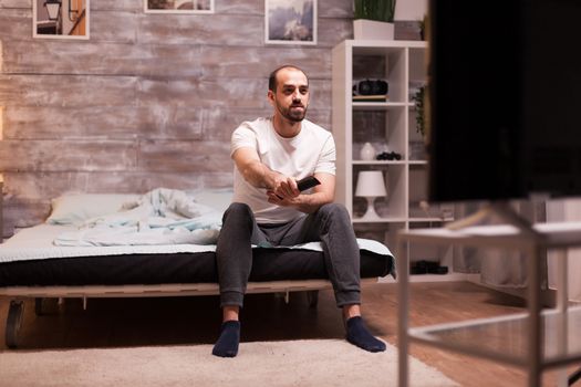 Relaxed man watching tv at night from the edge of his comfortable bed.