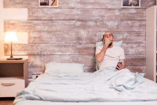 Tired man yawning at night in comfortable bed and holding smartphone.
