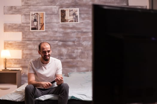 Cheerful man holding remote control watching tv at night.