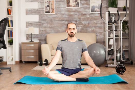 Man wearing sportswear while doing lotus yoga pose on mat in home and swiss ball in the background in time of covid-19 isolation.