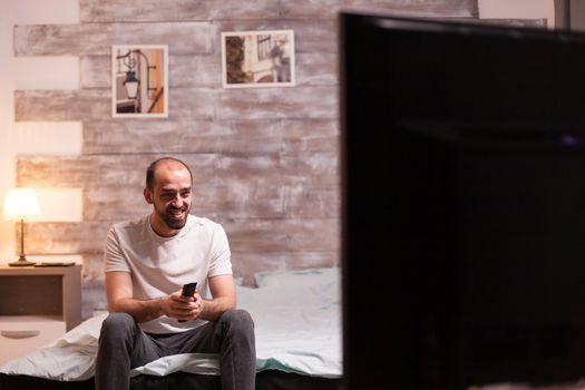 Excited man in pajamas watching tv at night in bedroom.