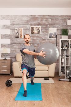 Fit man doing lunges on yoga mat using swiss ball during global pandemic.