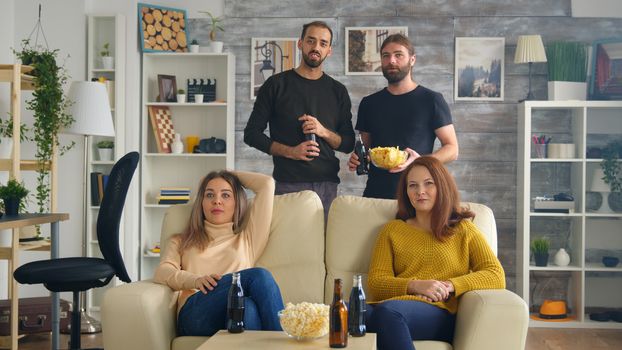 Group of friends watching a football match in living room drinking beer and eating snacks.