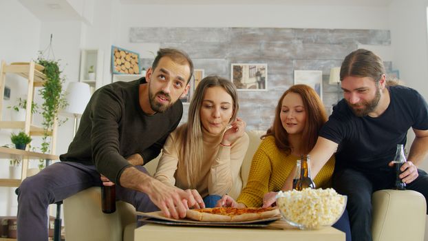 Close friends watches tv in living room cheering up while watching a football match on the couch. Friends eating snacks.