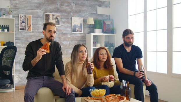 Young man eating chips while playing video games on tv with his friends sitting on the couch. Girl holding a beer bottle.