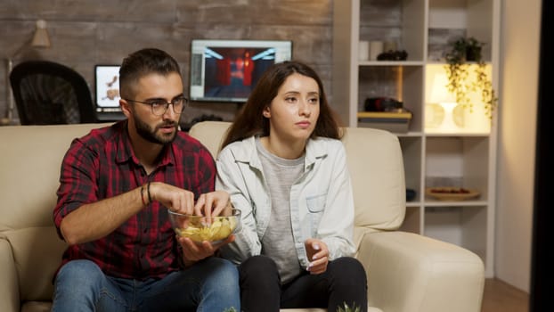 Caucasian young couple eating chips while watching television. Couple looking concerned at television.