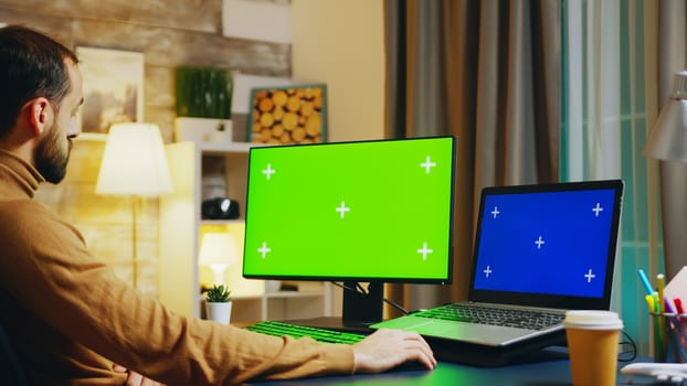 Zoom in shot of engineer at this desk working on computer with green screen. In the office at night.