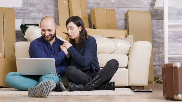 Couple smiling while sitting on the floor of their new apartment and doing online shopping using laptop.